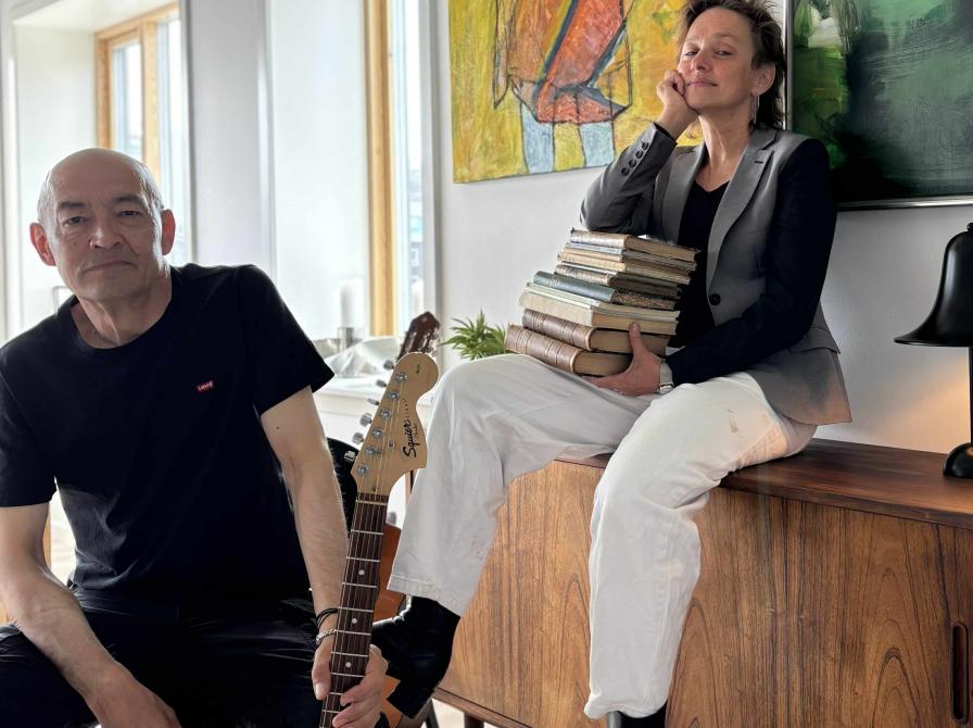 En mans i sort tøj sidder med en elguitar på en stol. Ved siden af sidder en kvinde med kort hår og hvide bukser på en skænk. Hun har en stak bøger i favnen.